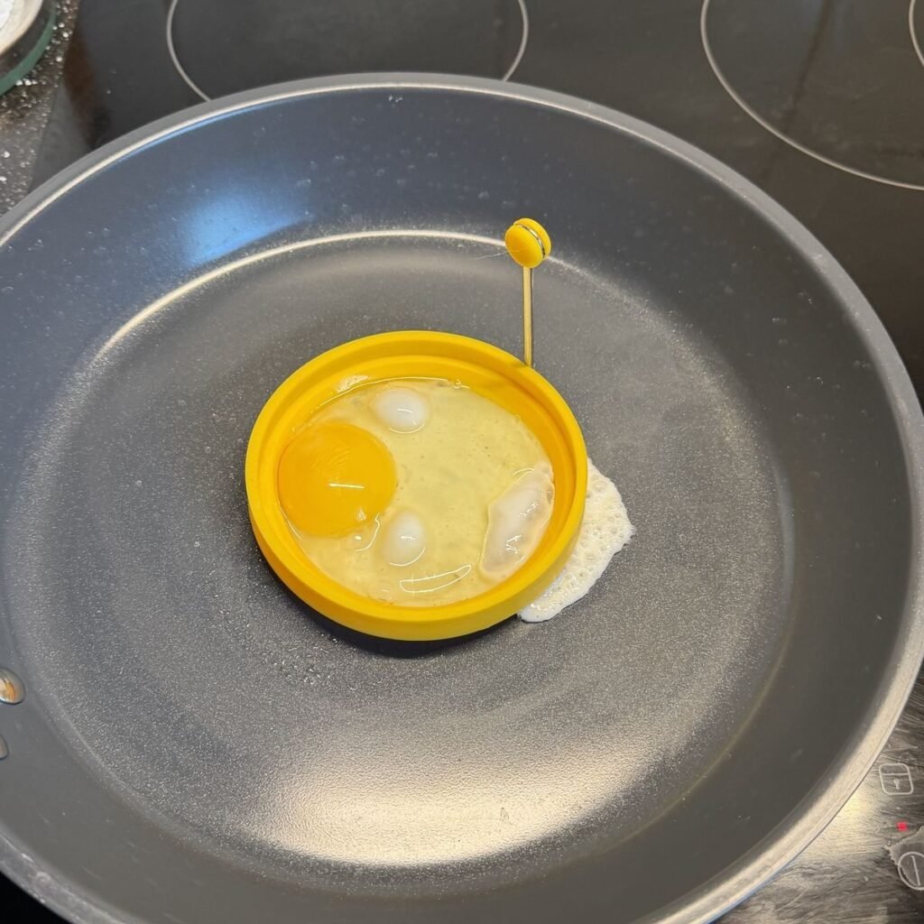 A yellow silicone egg ring on a non-stick pan with raw egg inside and egg white leaking out underneath