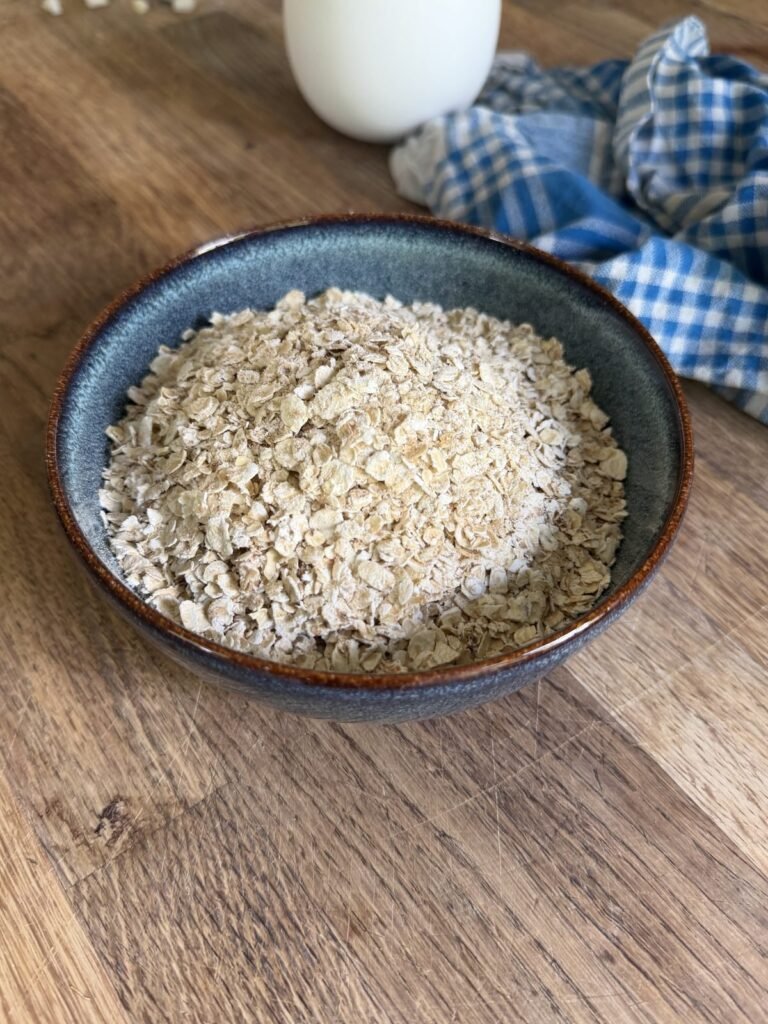 A bowl of rolled oats on a wooden table, with a jug of milk and a blue checked cloth behind.